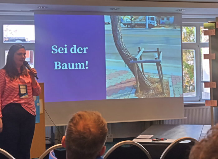 Eine Frau spricht in ein Mikrofon. Daneben auf der Folie ist ein Bild eines Baumes, der außerhalb einer Begrenzung wächst. Daneben steht: Sei der Baum!